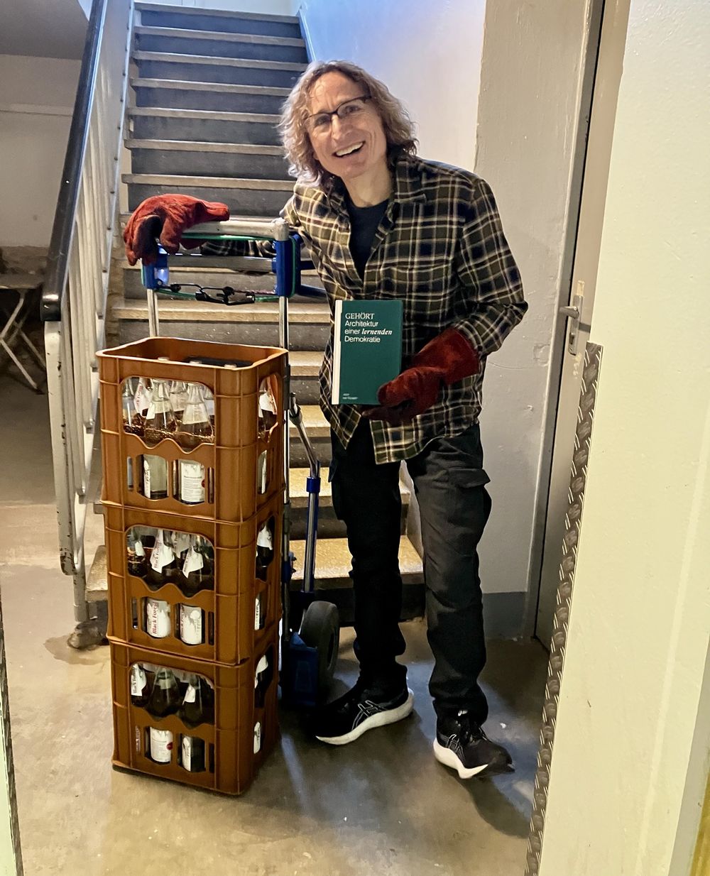 Jens van Rickeln im Treppenhaus mit Sackkarre, drei Getränkekisten und dem Buch GEHÖRT in der Hand.